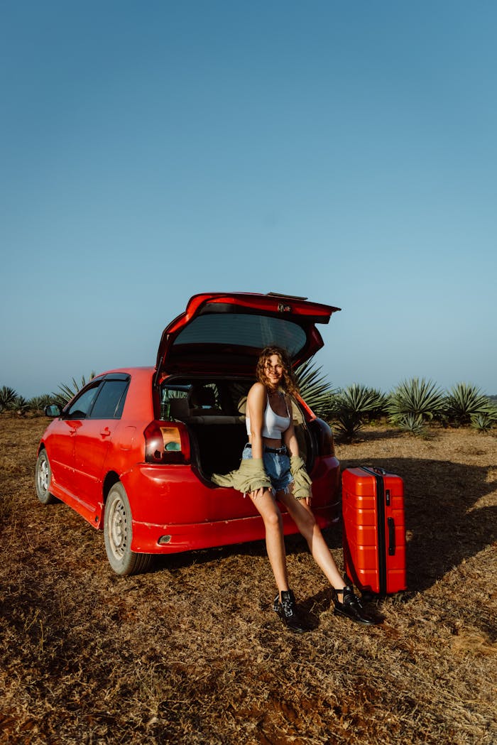 Young woman in denim shorts posing with suitcase beside a red car in a field.