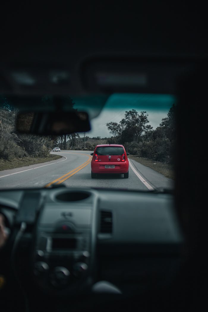 A red car driving on a winding countryside road, viewed from inside another vehicle.